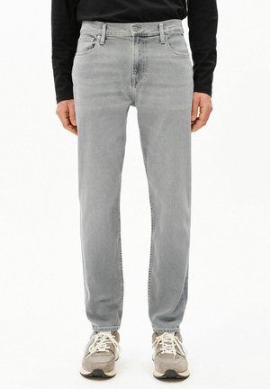 Personne portant un jean gris clair à jambes droites et des baskets beige, debout devant un fond blanc.
