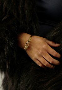 Guldkedja med ett länkat mönster, vilande på en hand; svart, fluffig tyg i bakgrunden. Slät yta, enkel och elegant form.