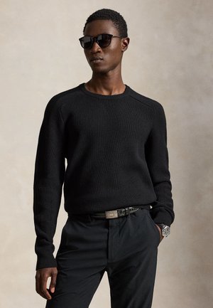 Homme portant un pull en maille noir, un pantalon noir, des lunettes de soleil noires, une ceinture noire et une montre argentée, debout devant un fond neutre.