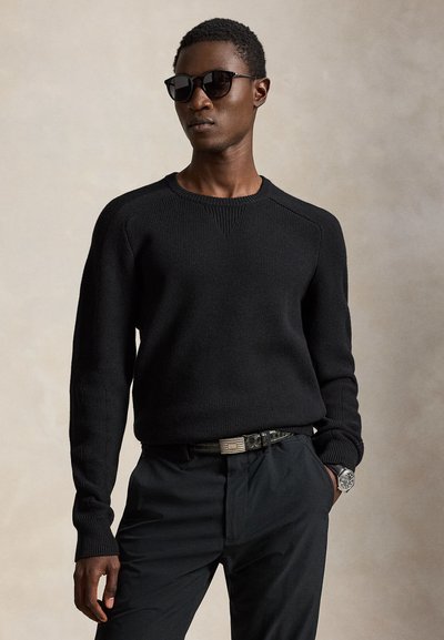 Homme portant un pull en maille noir, un pantalon noir, des lunettes de soleil noires, une ceinture noire et une montre argentée, debout devant un fond neutre.