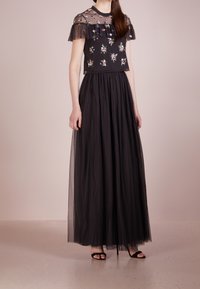 Robe maxi noire avec un bustier floral brodé transparent, des manches volantes et une jupe en tulle superposée à motifs à pois. Sandales noires à lanières.