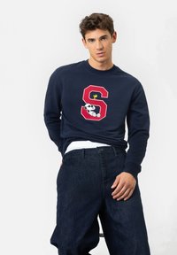 Jeune homme portant un sweat-shirt bleu marine avec une lettre "S" rouge mettant en vedette Snoopy et Woodstock, associé à un jean ample sombre, devant un fond blanc uni.