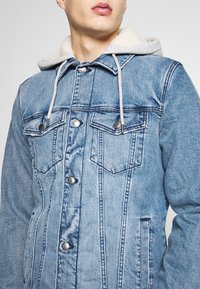 Veste en jean bleu clair avec une capuche en polaire, des boutons-pression en métal et deux poches poitrine, au design classique et avec des coutures contrastantes.