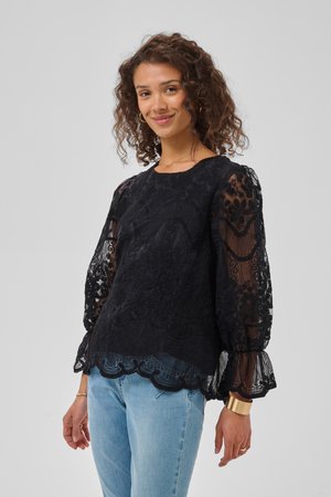Femme aux cheveux bouclés portant un chemisier en dentelle noire avec des manches bouffantes transparentes et un jean bleu clair, souriante sur un fond gris uni.