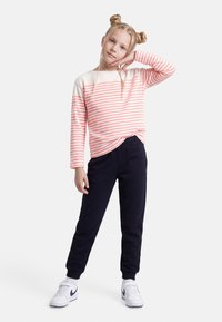 Κορίτσι με ξανθά μαλλιά σε δύο κότσους και κοτσίδες, φοράει ροζ και λευκή ριγέ μακρυμάνικη μπλούζα, μαύρο παντελόνι και λευκά αθλητικά παπούτσια, ποζάροντας με το χέρι στο πρόσωπο.