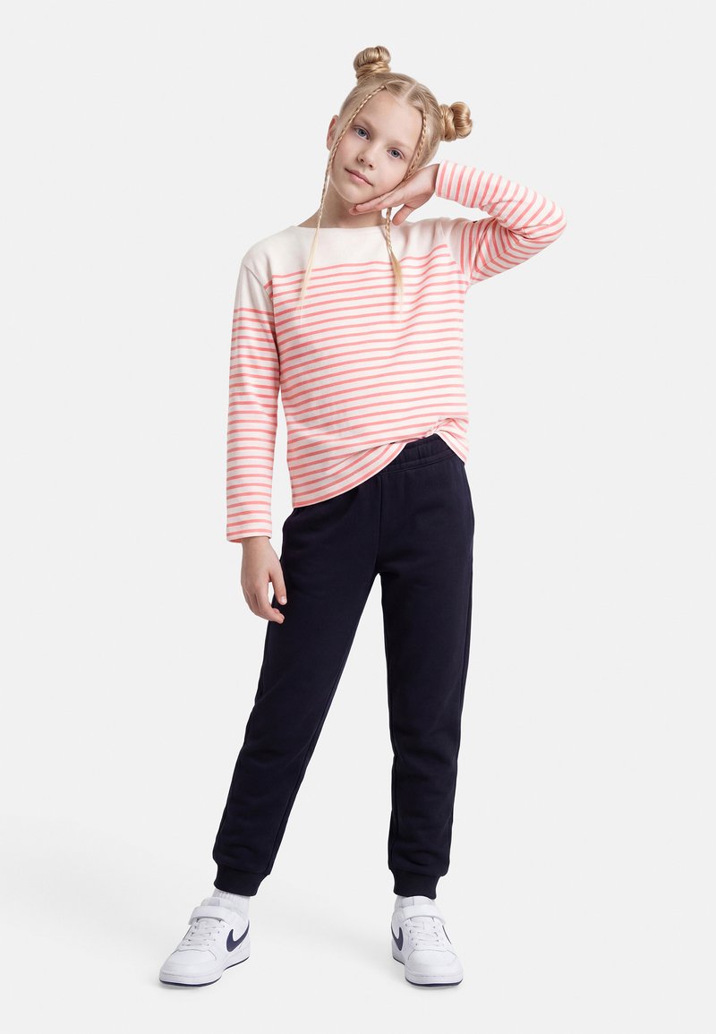 Κορίτσι με ξανθά μαλλιά σε δύο κότσους και κοτσίδες, φοράει ροζ και λευκή ριγέ μακρυμάνικη μπλούζα, μαύρο παντελόνι και λευκά αθλητικά παπούτσια, ποζάροντας με το χέρι στο πρόσωπο.