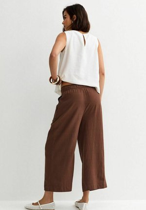 Donna con capelli corti e scuri, che indossa una canotta bianca senza maniche, pantaloni marroni a gamba larga, ballerine bianche traforate e braccialetti d'oro, in piedi contro una parete bianca.