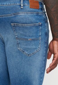 Jeans din denim albastru deschis cu o etichetă din piele la talie, un buzunar din spate cu un emblema colorată și detalii de cusătură standard.