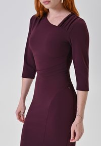 Bordeaux aansluitende jurk met driekwart mouwen, zijschoof en een decoratieve metalen accent bij de heup. Glad, rekbaar materiaal.