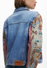 Jeansjack met een blauwe body, kleurrijk patroon op de mouwen en een bruine leren label aan de achterkant, gelabeld "Desigual."