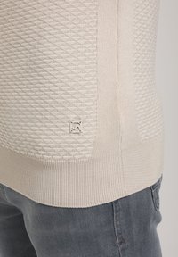 Hellbeige Strickpullover mit einem strukturierten Diamantmuster, geripptem Saum und einem kleinen Logo-Detail an der Seite. Weiches, leichtes Material.