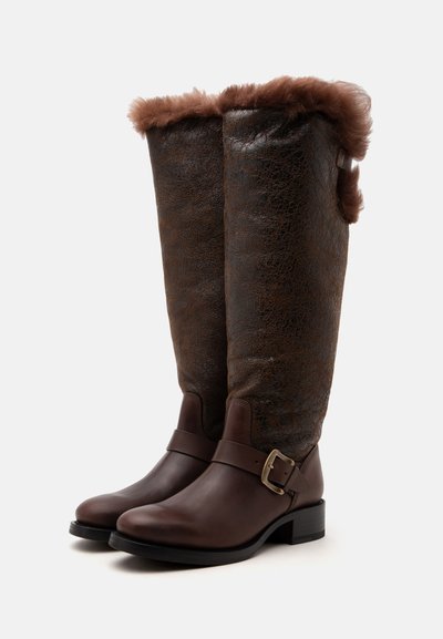 Paire de bottes en cuir marron montant jusqu'au genou avec bordure en fourrure en haut et sangle décorative avec boucle autour de la cheville sur fond blanc.