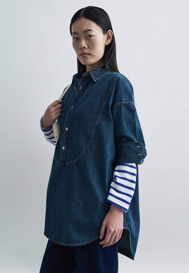 Τζιν πουκάμισο με εμπρόσθια κλεισίματα με κουμπιά, μεγάλο γιακά και κυρτή λήξη. Διαθέτει μανίκια που έχουν γυριστεί πάνω από μια ριγωτή μπλούζα μακριάς μανίκας. Σκούρο μπλε χρώμα.