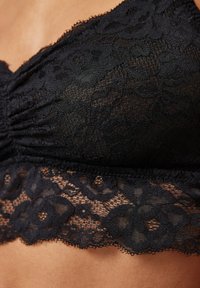 Bralette en dentelle noire avec un motif floral, présentant un tissu doux, des détails froncés et des bords festonnés le long du bas.