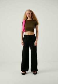 Jauna sieviete ar viļņainiem blondiem matiem, kas valkā rozā saulesbrilles, tumši zaļu īsu T-kreklu, melnas plašas bikses, melnas sandales un rozā pleca somu.