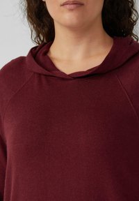 Burgundowa bluza z kapturem o miękkiej teksturze, z prążkowanym dekoltem i rękawami raglanowymi. Projekt jest prosty, bez widocznych wzorów lub akcentów.