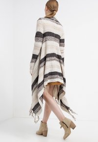Mujer con una coleta castaña clara, vistiendo un cárdigan largo de punto a rayas en tonos beige y marrón, una falda color tostado y botines de ante beige con tacones cuadrados.