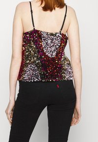 Top multicolore con paillettes e spalline nere regolabili, caratterizzato da una superficie scintillante e testurizzata con un motivo intricato. Indossato con jeans neri.