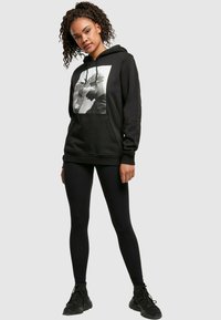Sudadera negra con un estampado gráfico en la parte frontal, que presenta un retrato en blanco. Tejido suave, bolsillo tipo canguro, puños acanalados. Llevada con leggings negros.