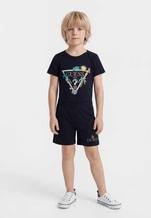 Garçon blond debout, portant un t-shirt d'été noir Guess et un short assorti avec des baskets blanches sur un fond blanc uni.