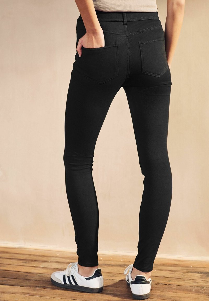 Personne portant un jean skinny noir et des baskets blanches, debout avec une main dans la poche arrière sur un sol en bois contre un mur beige.
