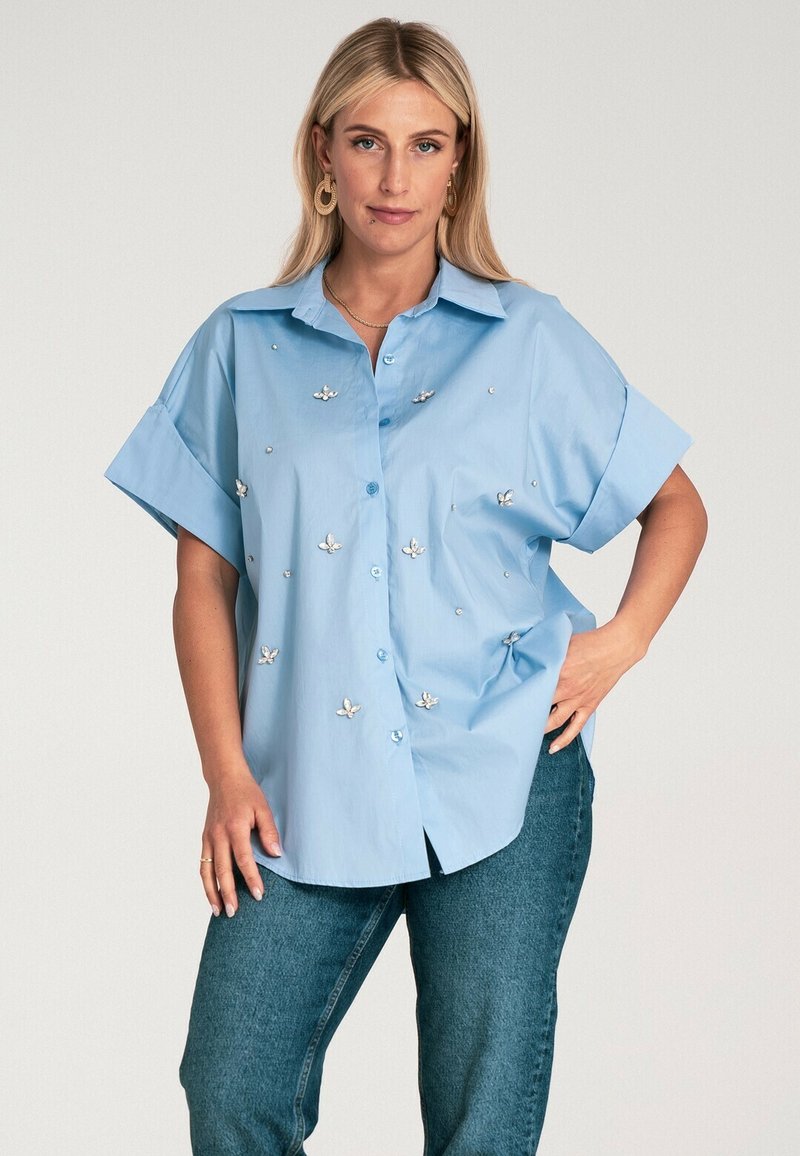 Jasnoniebieska koszula z krótłym rękawem i kołnierzykiem, ozdobiona kwiatowymi wzorami i małymi dekoracyjnymi kamieniami. Noszona z dżinsami.