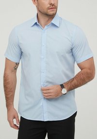 Chemise légère bleue à manches courtes avec fermeture à boutons, col et texture lisse. Portée avec une montre au poignet, dotée d'un bracelet noir et d'un cadran blanc.