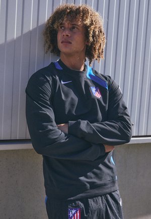 Black long-sleeve athletic sweatshirt with blue accents and a shield logo. The model has curly hair and is wearing matching black pants.