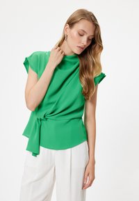 Blusa leggera verde con un colletto alto, maniche corte e un dettaglio con nodo in vita. Indossata con pantaloni bianchi a gamba larga.