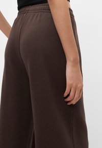 Καφέ, ψηλόμεσες παντελόνες με φαρδύ πόδι, με λεία υφή, ελαστική μέση και ορατές λεπτομέρειες στις ραφές στο  πλάι. Χέρι που στηρίζεται κοντά στο ύφασμα.