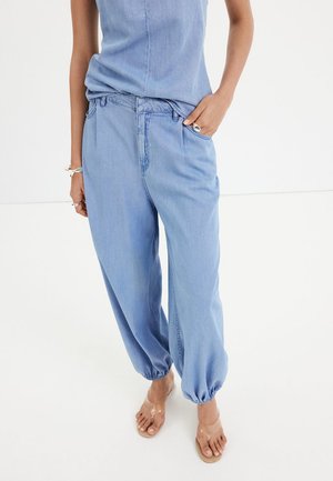Mujer vistiendo pantalones jogger de denim azul claro con puños elásticos y una camiseta sin mangas a juego, de pie descalza sobre un suelo blanco.