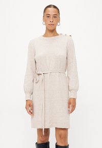 Gebreide beige sweater jurk met lange mouwen, een riem rond de taille en een gouden knoopdetail op de schouder. Zachte textuur en relaxte pasvorm.