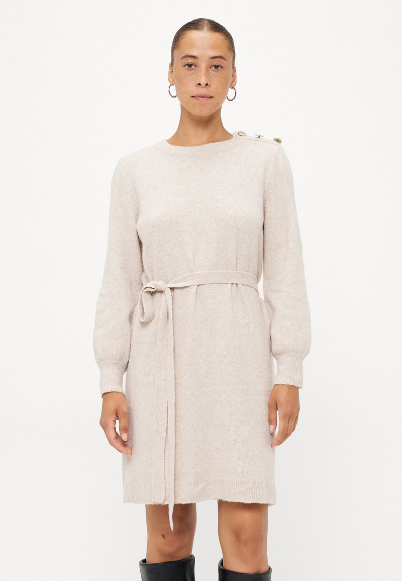 Gebreide beige sweater jurk met lange mouwen, een riem rond de taille en een gouden knoopdetail op de schouder. Zachte textuur en relaxte pasvorm.