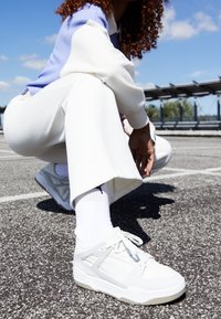 Baskets blanches avec des accents gris, tissu texturé et design structuré. Portées avec des chaussettes blanches et un pantalon blanc ample dans un cadre extérieur.