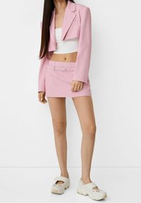 Blazer corto rosa abbinato a una mini gonna con cintura. Top corto bianco sotto. Tessuto leggero, texture liscia, abbinato a scarpe bianche.