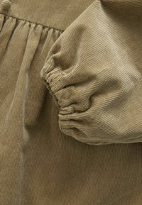 Tessuto di velluto a coste verde oliva con righe verticali, caratterizzato da polsini a manica arricciati e elementi di design plissettati. Morbida texture e forma strutturata.