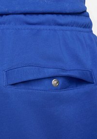 Blå bomullstyg med en bakficka, utrustad med en enkel klaff och en silverfärgad tryckknapp. Mjuk textur med jämn sömnad.