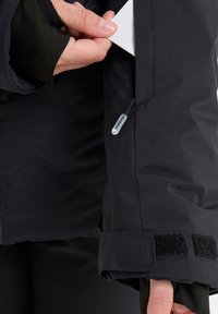 Black insulated jacket with a textured finish. Features a Velcro cuff and a small visible pocket on the sleeve, with a pull tab for access.