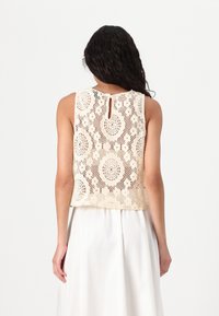 Lace Cropped Top i krem med sirkulære mønstre, med en nøkkelhullsåpning bak. Stoffet er lett med en teksturert overflate.