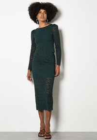 Vestido de encaje verde oscuro con mangas largas, silueta ajustada y dobladillo en forma de ola, que presenta un patrón floral texturizado. Lleva unos tacones negros con tiras.