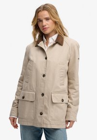 Mujer con chaqueta beige con cuello marrón y botones negros sobre camisa blanca y jeans azules, de pie frente a un fondo blanco liso.