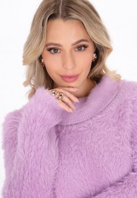 Helllila, flauschiger Pullover mit hohem Kragen und gerippten Details. Das Model hält eine Hand nah am Kinn, um Ringe und natürliche Nägel zu präsentieren.