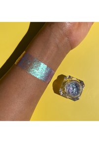 Iridescent cosmetische shimmer aangebracht op de huid, met een mengeling van blauwe, paarse en gouden tinten. De verpakking is een heldere, hoekige pot.