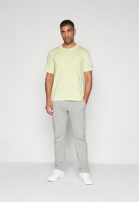 Helles gelbes T-Shirt mit kurzen Ärmeln und gesticktem "Champion"-Logo, kombiniert mit grauen Sweatpants mit Logo-Akzenten. Weiße Sneakers.