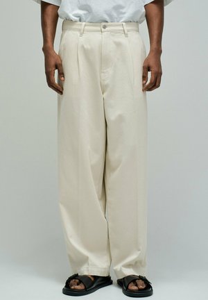 Homme portant un pantalon beige ample avec des plis devant et des sandales noires à enfiler, sur un fond clair uni.