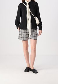Schwarze Bluse mit langen Ärmeln und Spitzenakzenten, kombiniert mit grau-schwarz karierten Shorts. Ein cremefarbener Schal und schwarze Mokassins runden das Outfit ab.