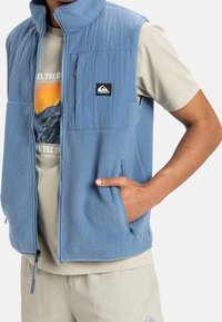 Světle modrá fleecová vesta s texturovaným předním panelem, vybavená kapsami na zip a nášivkou na hrudi, v kombinaci se světle barevnou košilí pod ní.