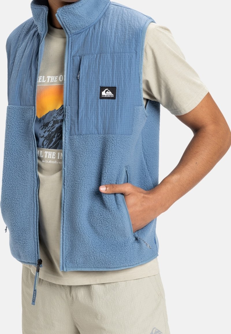 Světle modrá fleecová vesta s texturovaným předním panelem, vybavená kapsami na zip a nášivkou na hrudi, v kombinaci se světle barevnou košilí pod ní.