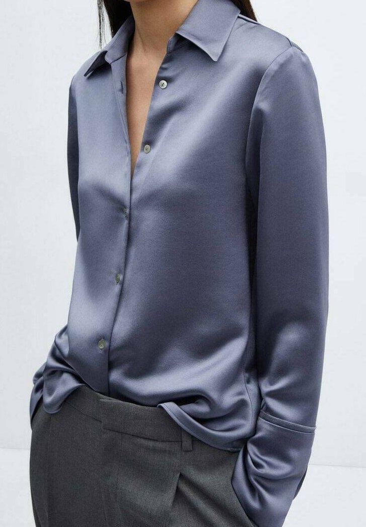 Femme portant une chemise boutonnée en satin bleu brillant à manches longues, rentrée dans un pantalon gris sur mesure.
