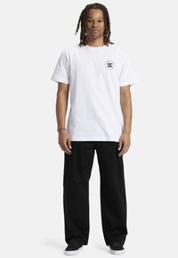 White cotton t-shirt with a black logo on the left chest, paired with loose-fitting black pants and black skate shoes.